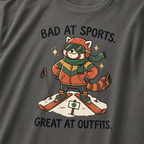 Close-up of a pepper gray t-shirt featuring a stylish red panda skier posing in coordinated winter gear with “Bad at Sports. Great at Outfits.” text.