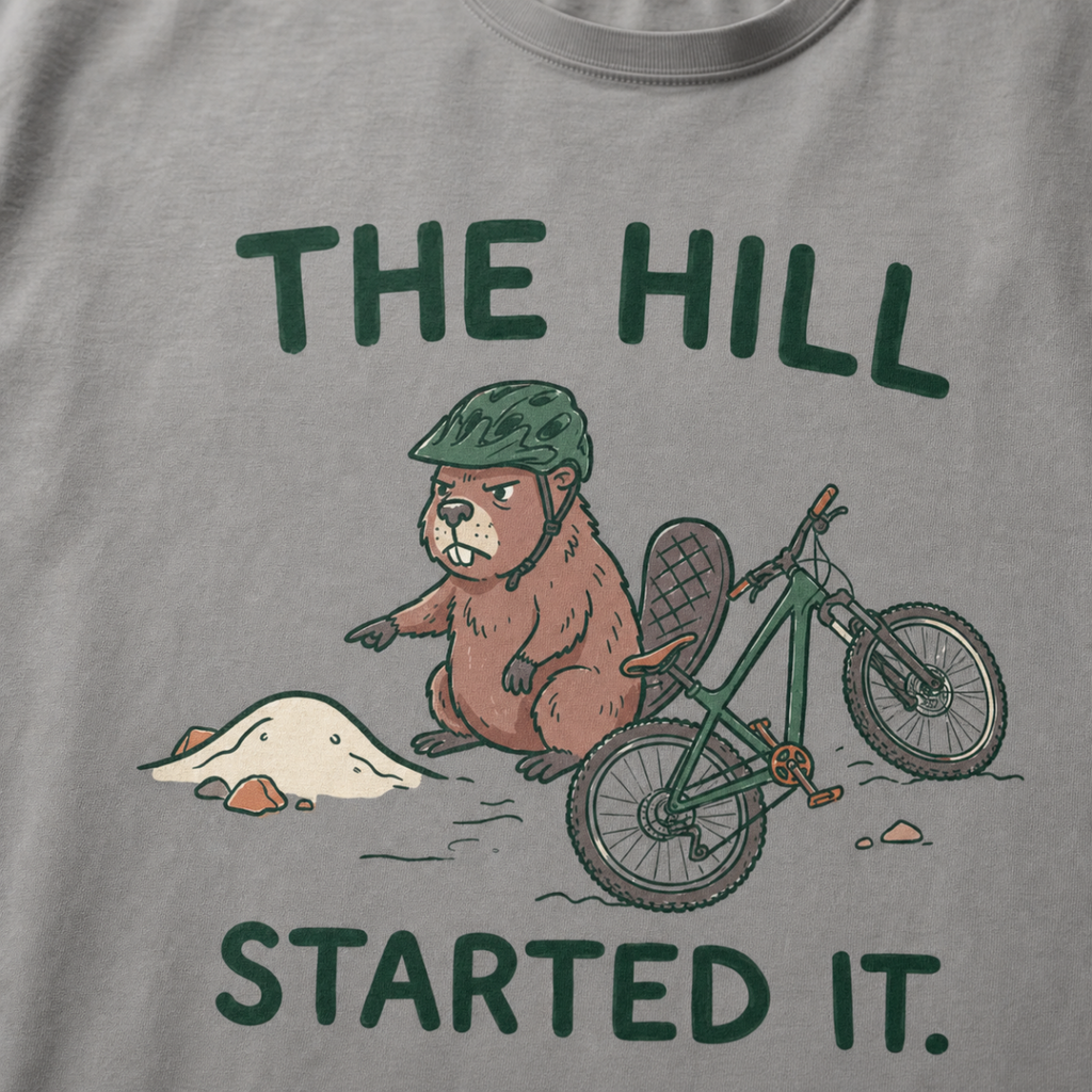 Close-up of a grey t-shirt featuring a grumpy beaver in a bike helmet pointing at a small dirt hill beside a mountain bike, with “The Hill Started It.” text.