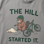 Close-up of a grey t-shirt featuring a grumpy beaver in a bike helmet pointing at a small dirt hill beside a mountain bike, with “The Hill Started It.” text.