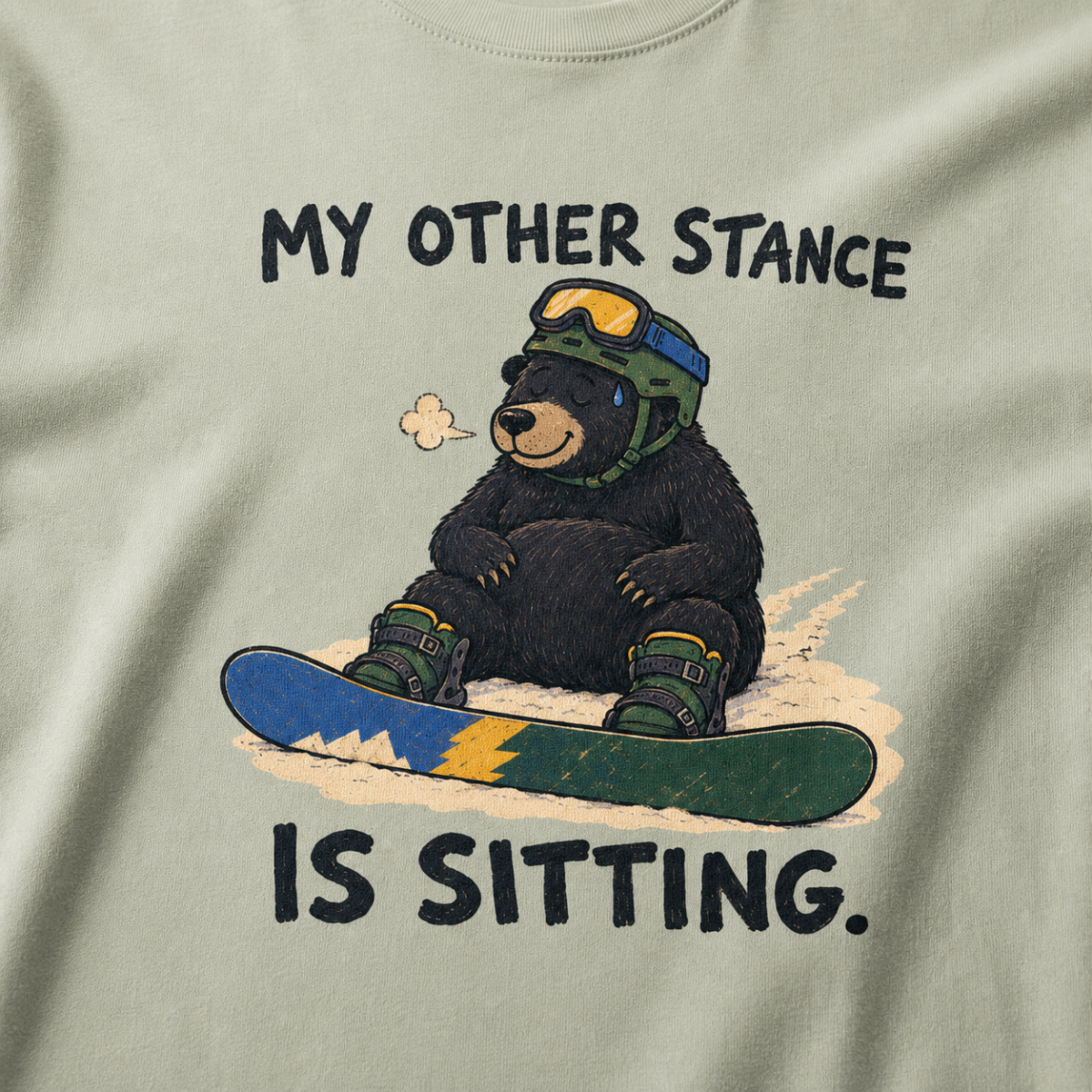 Close-up of a bay green t-shirt featuring a tired bear sitting in the snow beside a snowboard, with “My Other Stance Is Sitting.” text.