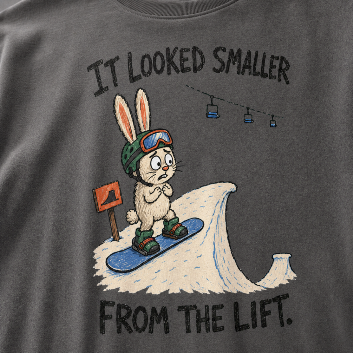 Close-up of a pepper gray t-shirt featuring a nervous rabbit snowboarder at the edge of a large snow jump, with “It Looked Smaller From the Lift.” text.