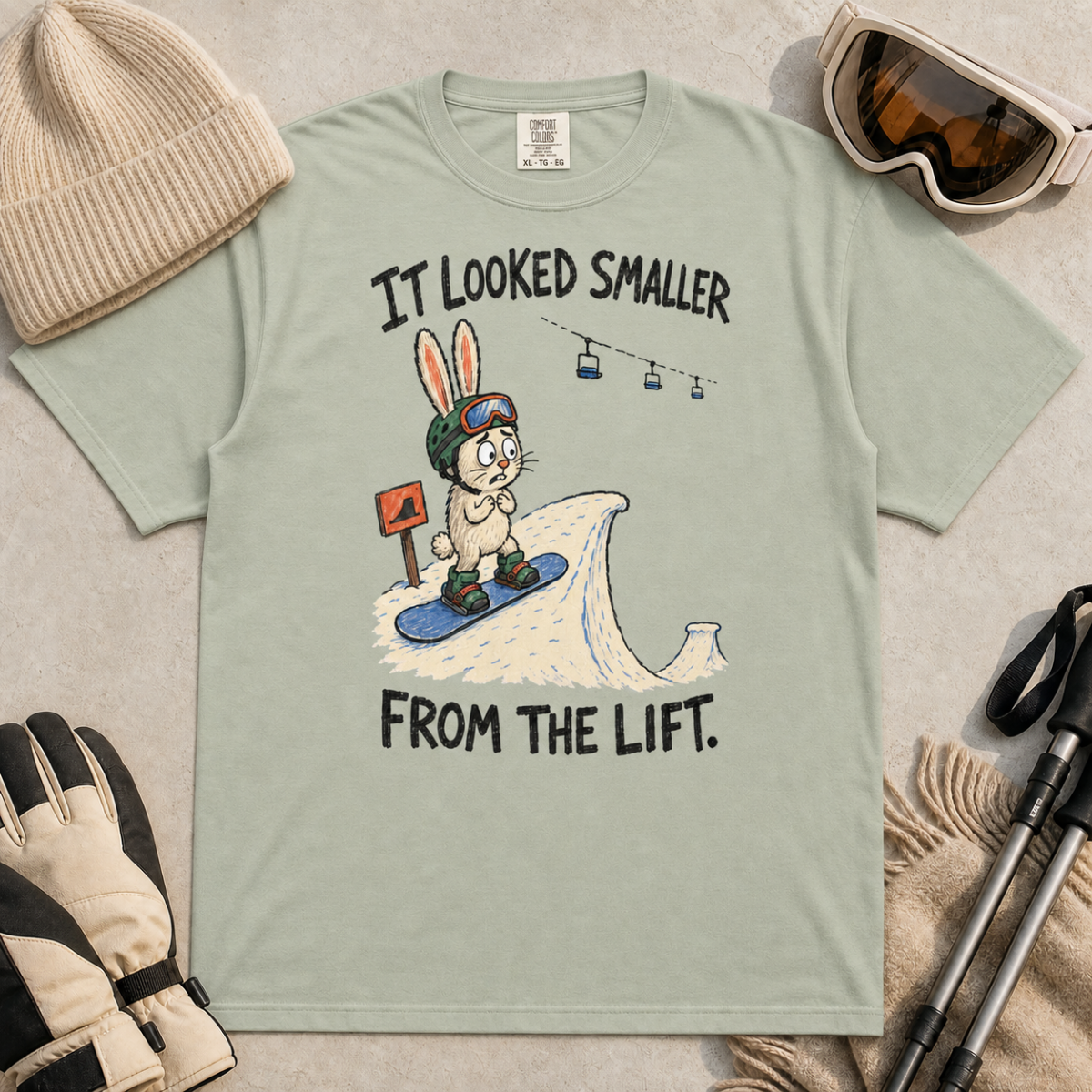 Flat lay of a bay green t-shirt with a humorous snowboarding design showing a worried rabbit facing an unexpectedly steep jump.