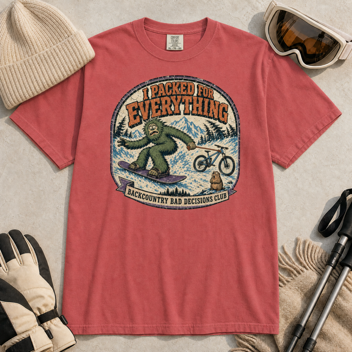 Crimson red t-shirt featuring a vintage backcountry badge design with Bigfoot snowboarding, a mountain bike, snowy peaks, and “I Packed for Everything” lettering.