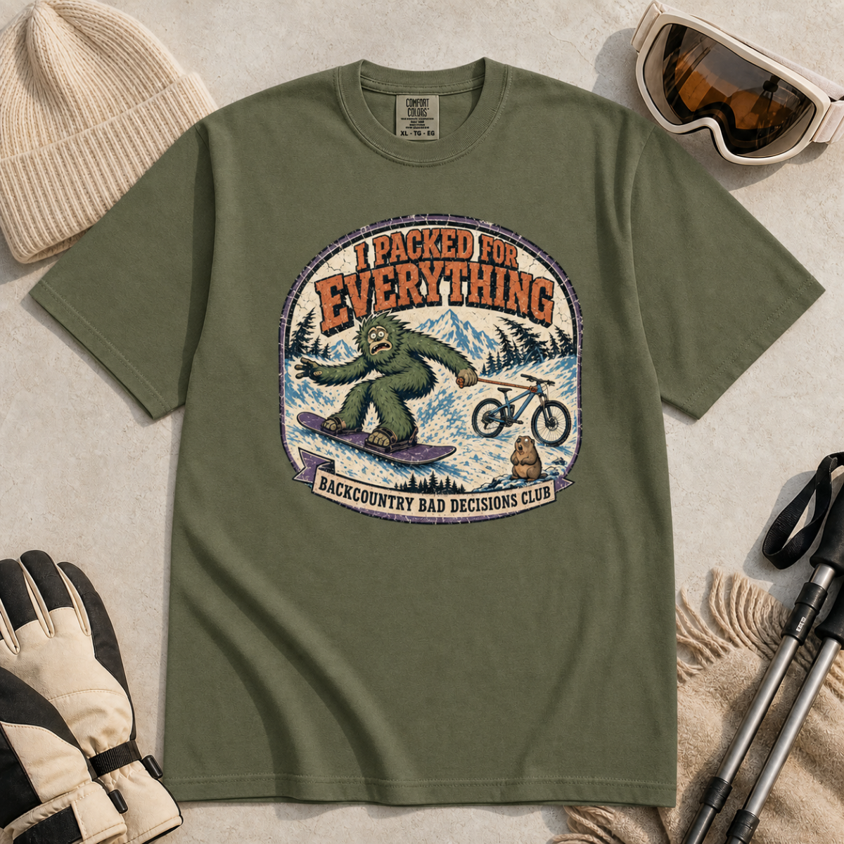 Hemp green t-shirt featuring a vintage backcountry badge design with Bigfoot snowboarding, a mountain bike, snowy peaks, and “I Packed for Everything” lettering.