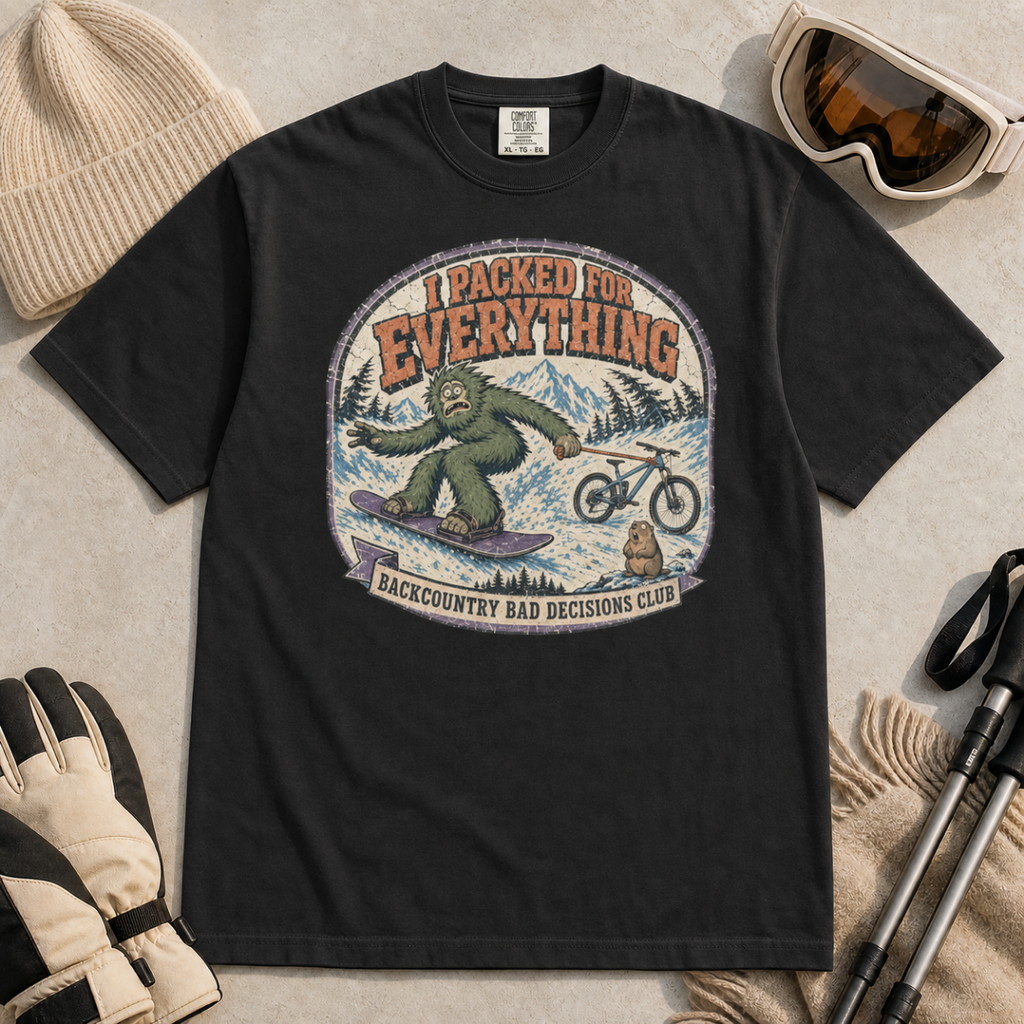 Black t-shirt featuring a vintage backcountry badge design with Bigfoot snowboarding, a mountain bike, snowy peaks, and “I Packed for Everything” lettering.