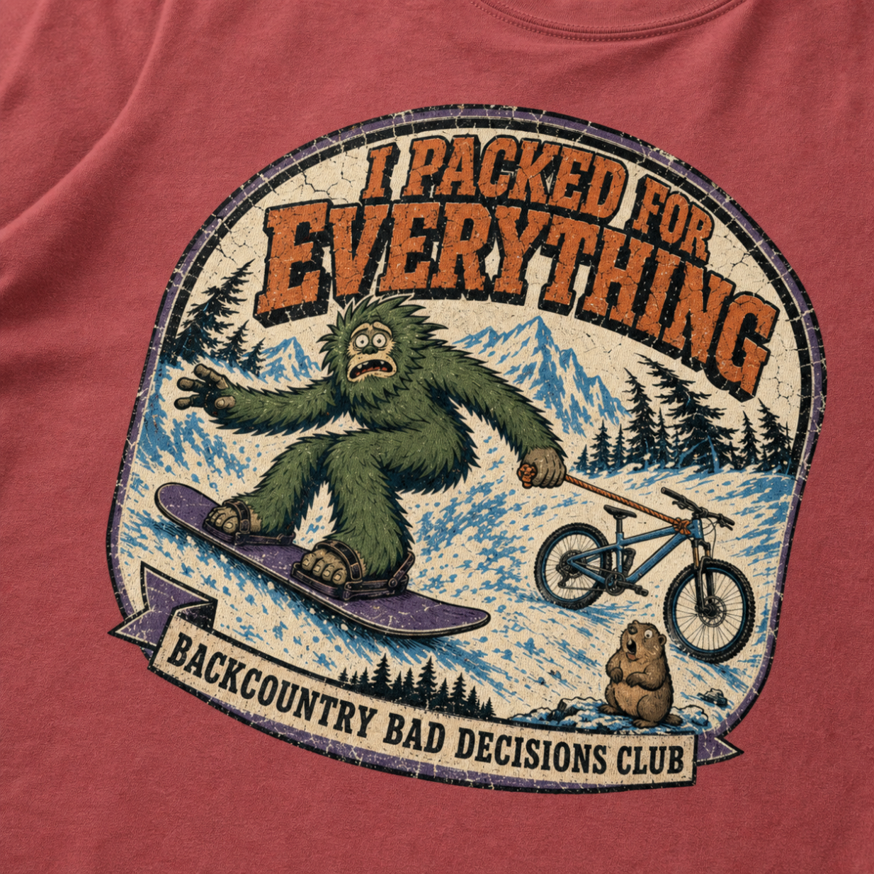 Close-up of a crimson red t-shirt featuring a distressed Bigfoot snowboarding while towing a mountain bike, with “I Packed for Everything” and “Backcountry Bad Decisions Club” text.