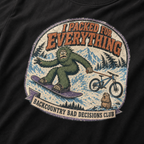 Close-up of a black t-shirt featuring a distressed Bigfoot snowboarding while towing a mountain bike, with “I Packed for Everything” and “Backcountry Bad Decisions Club” text.