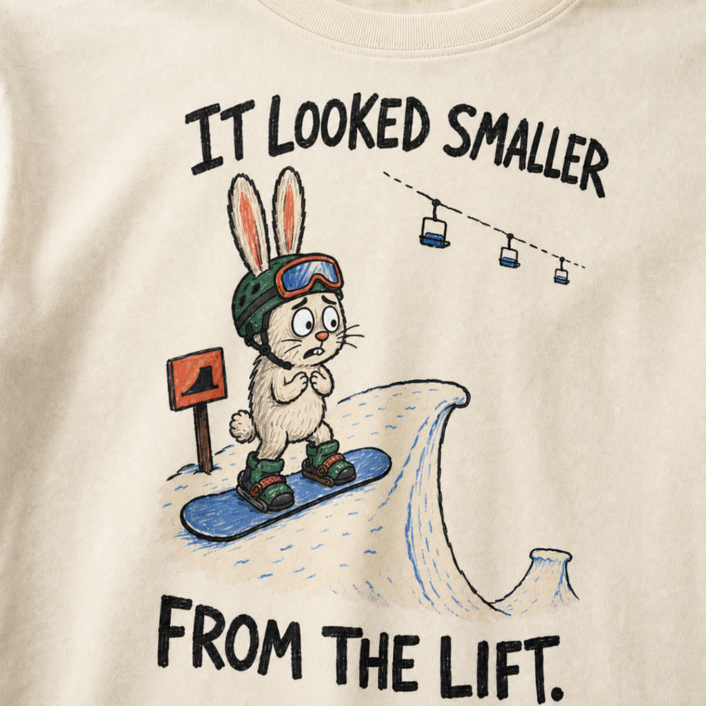 Close-up of an ivory white t-shirt featuring a nervous rabbit snowboarder at the edge of a large snow jump, with “It Looked Smaller From the Lift.” text.