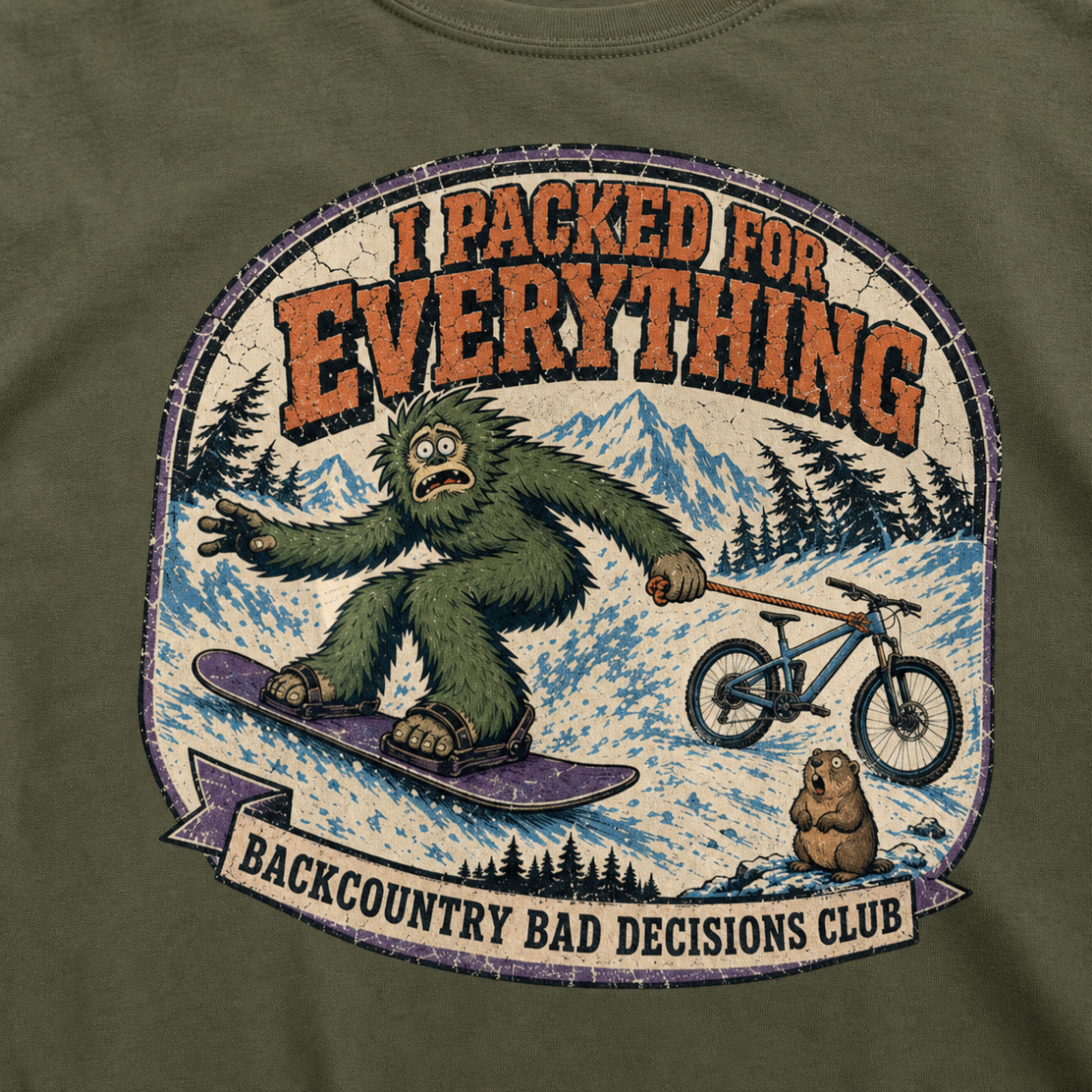 Close-up of a hemp green t-shirt featuring a distressed Bigfoot snowboarding while towing a mountain bike, with “I Packed for Everything” and “Backcountry Bad Decisions Club” text.
