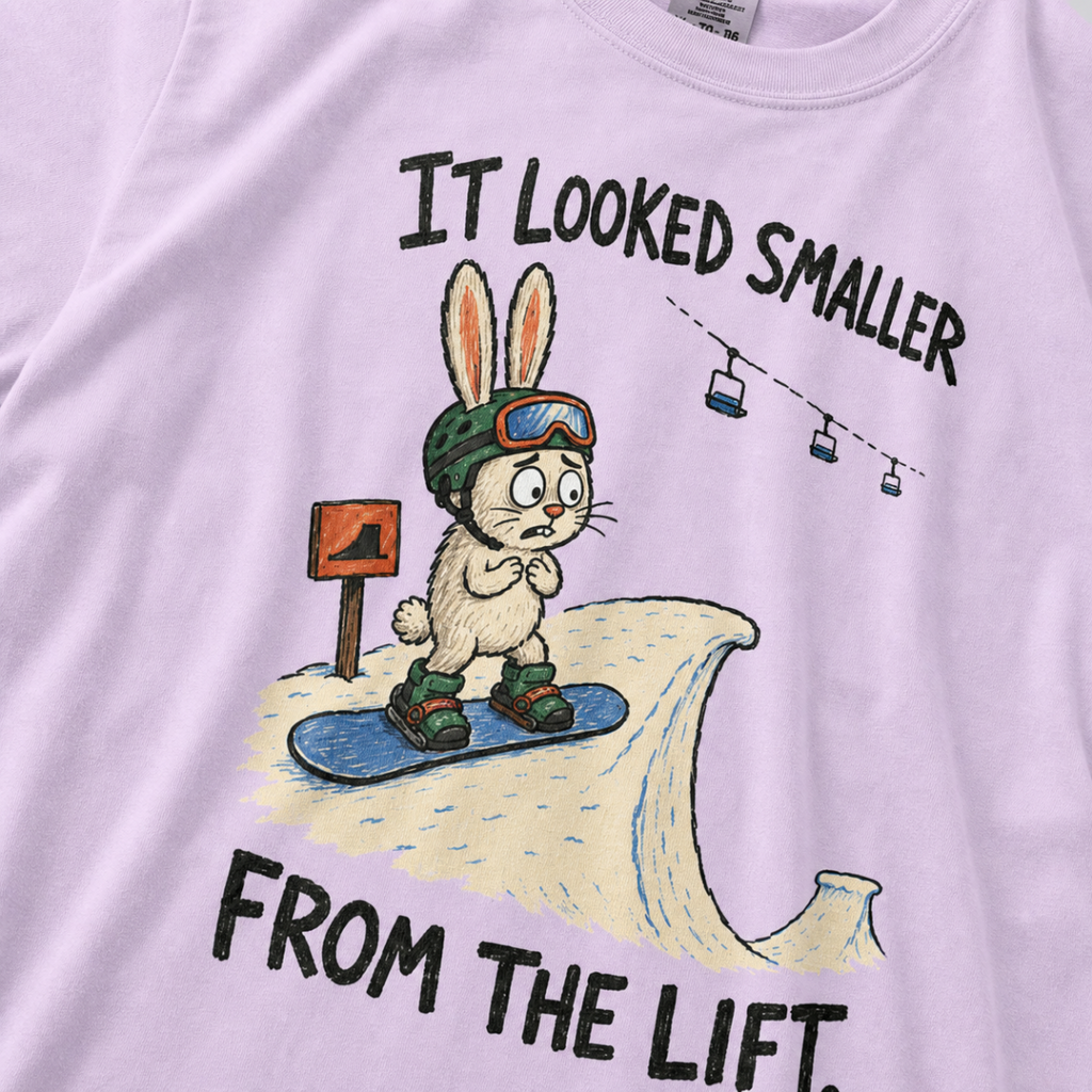 Close-up of an orchid t-shirt featuring a nervous rabbit snowboarder at the edge of a large snow jump, with “It Looked Smaller From the Lift.” text.