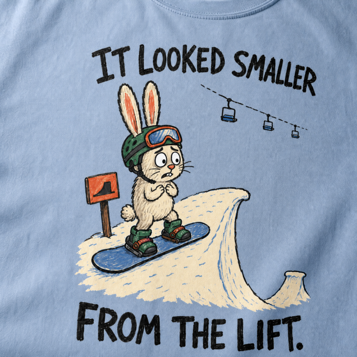 Close-up of a washed denim blue t-shirt featuring a nervous rabbit snowboarder at the edge of a large snow jump, with “It Looked Smaller From the Lift.” text.