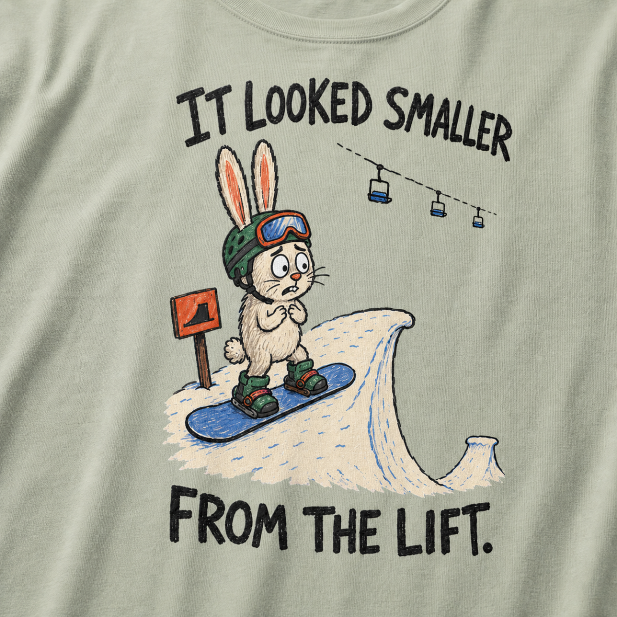 Close-up of a bay green t-shirt featuring a nervous rabbit snowboarder at the edge of a large snow jump, with “It Looked Smaller From the Lift.” text.