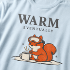 Close-up of a chambray blue t-shirt design showing a ski-goggle-wearing squirrel beside a hot drink under “Warm Eventually” text.