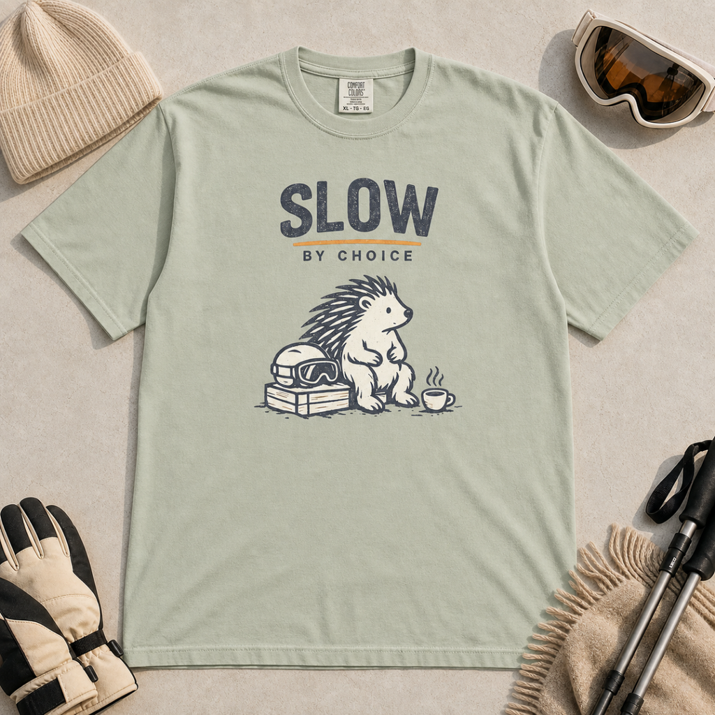 Bay green t-shirt featuring a seated hedgehog beside ski goggles and a hot drink beneath “Slow by Choice” text.