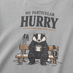 Close-up of a granite gray t-shirt featuring a badger seated on a snowy bench with a mug, ski poles and goggles beneath the words “No Particular Hurry.”