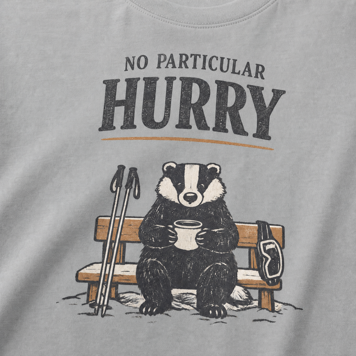 Close-up of a granite gray t-shirt featuring a badger seated on a snowy bench with a mug, ski poles and goggles beneath the words “No Particular Hurry.”