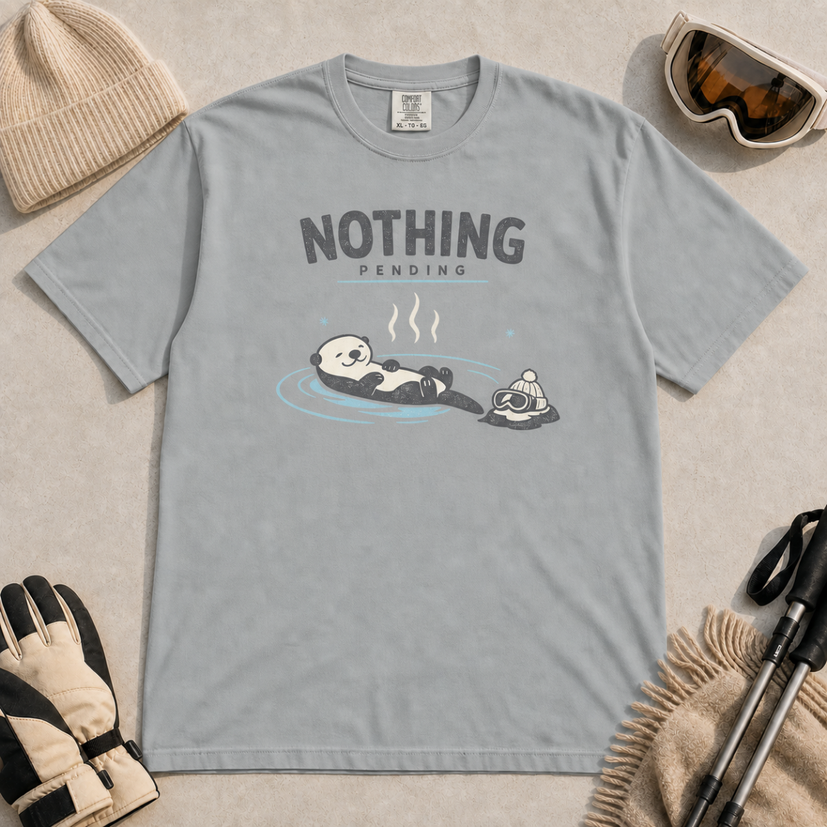 Granite gray t-shirt featuring a relaxed otter floating in a hot spring beside winter goggles and a beanie, with the words “Nothing Pending.”