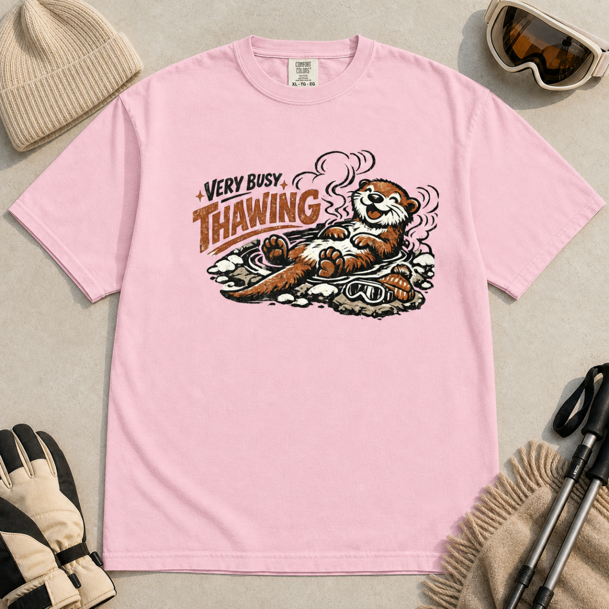Blossom pink t-shirt featuring a smiling otter thawing in a rocky hot spring beside ski goggles and a beanie.