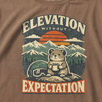 Close-up of an espresso brown t-shirt design featuring a small mountain rodent holding a mug beside ski goggles, with sunset peaks and the phrase “Elevation Without Expectation.”