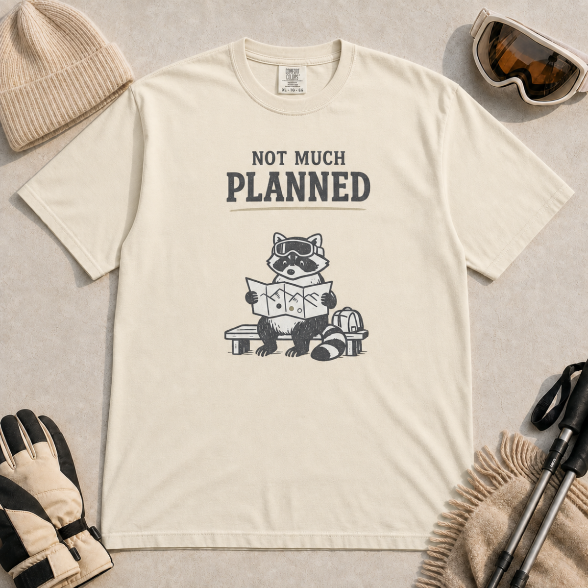 Ivory white t-shirt featuring a raccoon in ski goggles sitting on a bench and studying a trail map, with the words “Not Much Planned.”