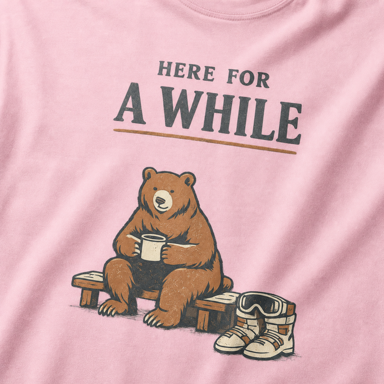 Close-up of a blossom pink t-shirt featuring a bear holding a mug beside ski boots and goggles with the phrase “Here for a While.”
