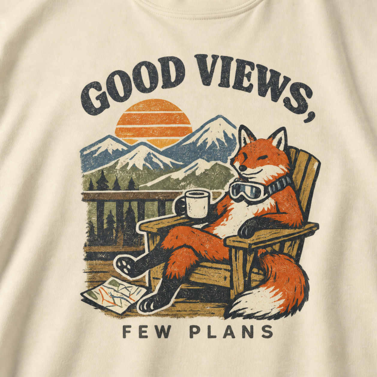 Close-up of an ivory white t-shirt design showing a fox relaxing with a mug before mountain scenery and the phrase “Good Views, Few Plans.”