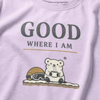 Close-up of an orchid purple t-shirt featuring a small white bear holding a warm drink beside ski goggles with the phrase “Good Where I Am.”