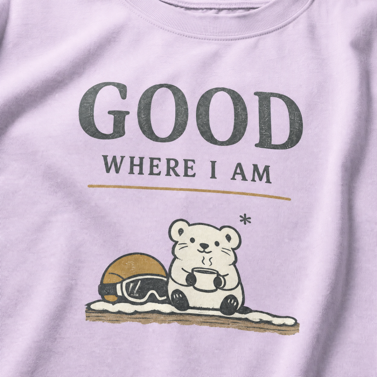 Close-up of an orchid purple t-shirt featuring a small white bear holding a warm drink beside ski goggles with the phrase “Good Where I Am.”