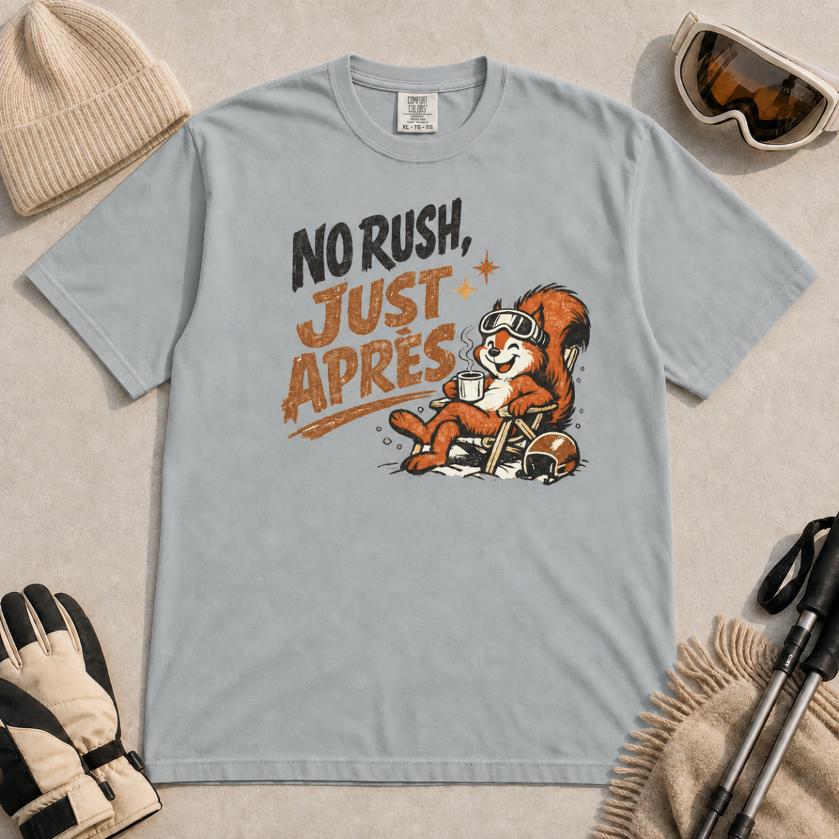 Granite gray t-shirt featuring a relaxed red squirrel in ski goggles sitting in a chair with a steaming mug and ski helmet, beside the words “No Rush, Just Après.”