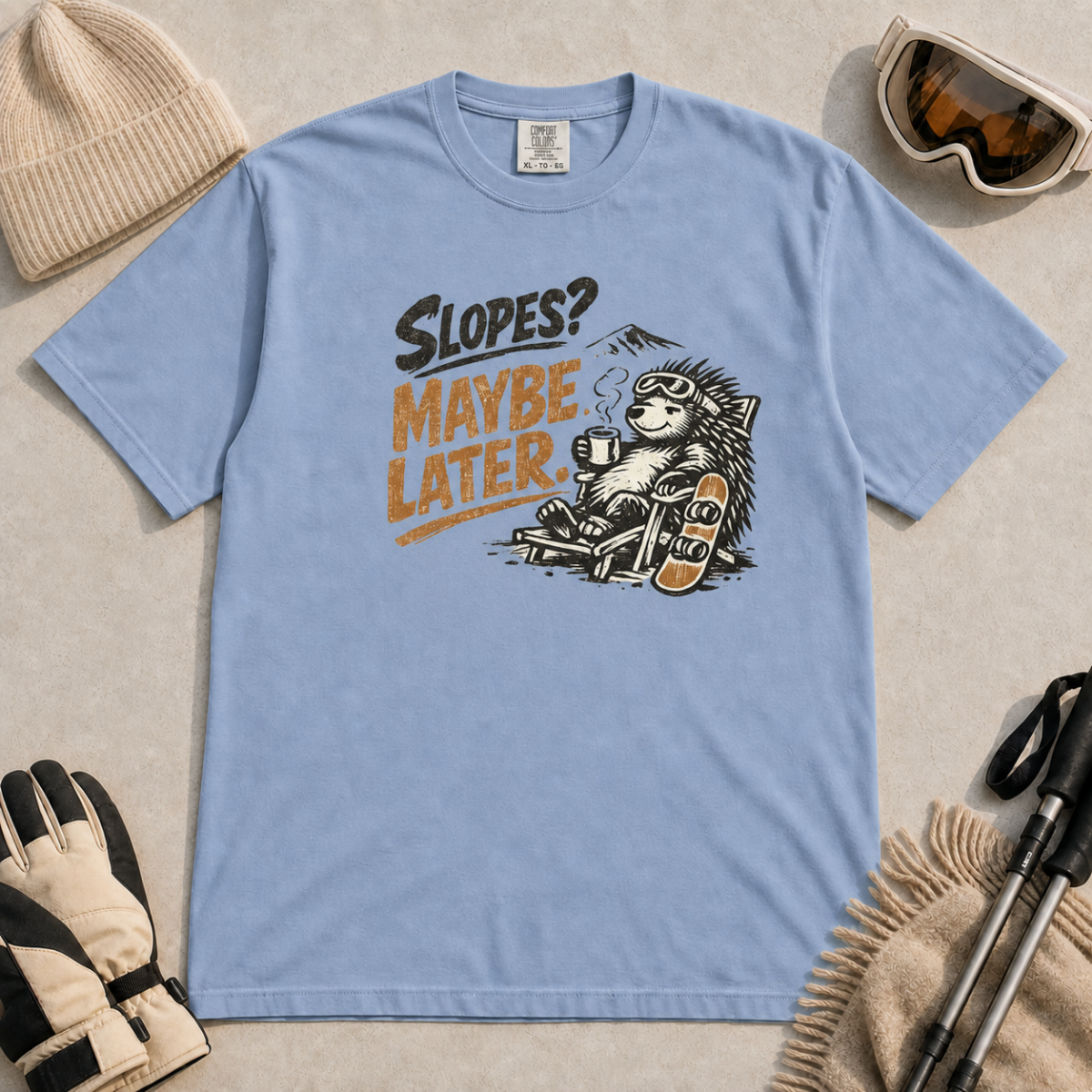 Washed denim blue t-shirt featuring a relaxed hedgehog in a chair with a hot drink and snowboard beneath “Slopes? Maybe Later.” text.
