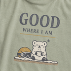Close-up of a bay green t-shirt featuring a small white bear holding a warm drink beside ski goggles with the phrase “Good Where I Am.”