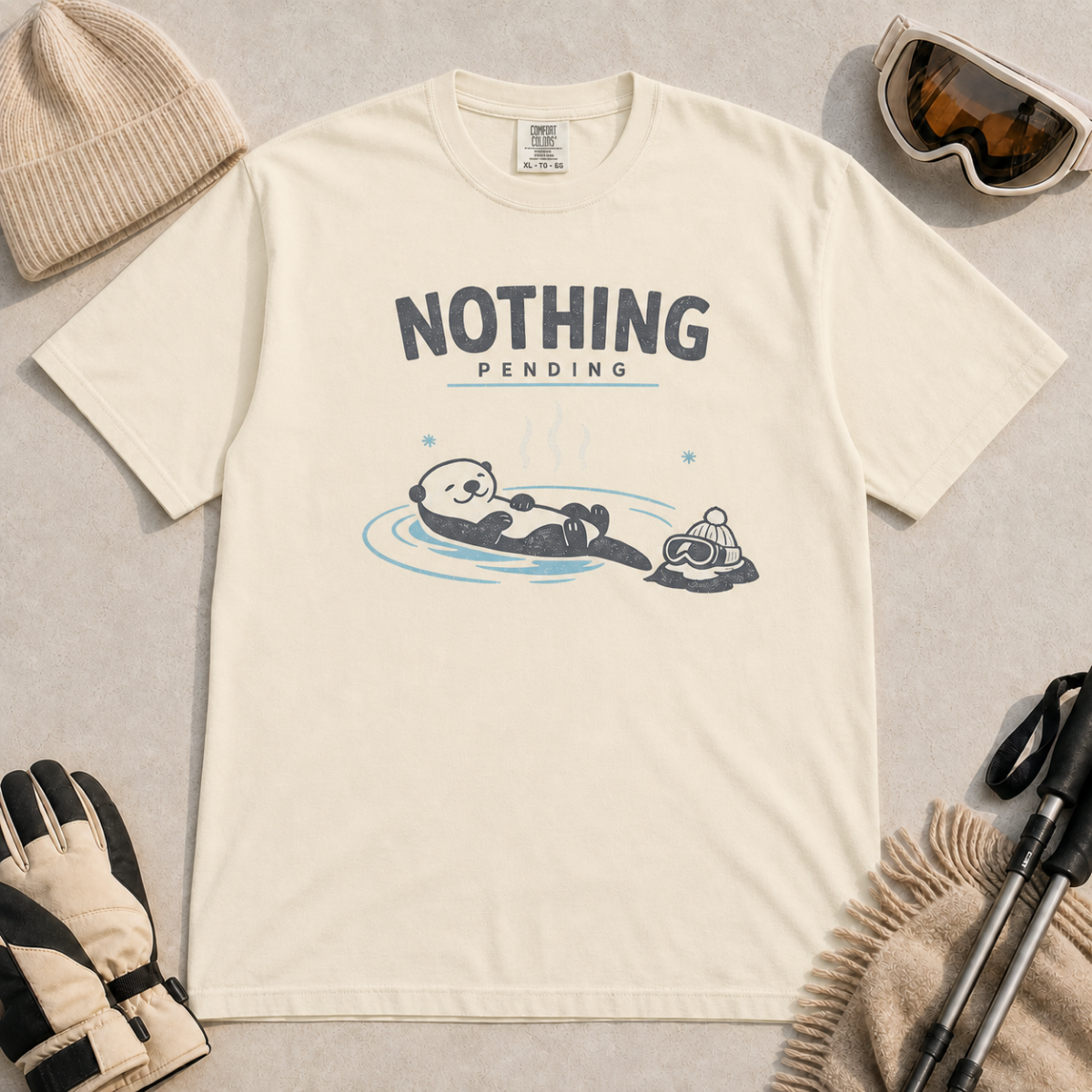 Ivory white t-shirt featuring a relaxed otter floating in a hot spring beside winter goggles and a beanie, with the words “Nothing Pending.”