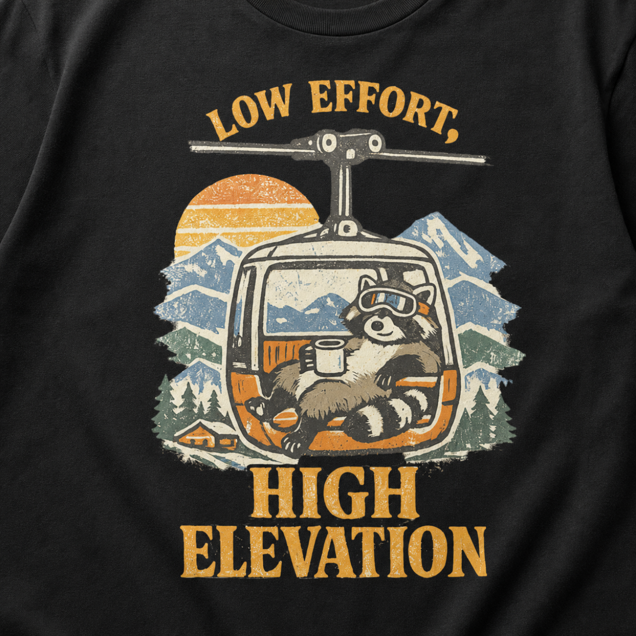 Close-up of a vintage raccoon gondola design with snowy mountains, a sunset and the words “Low Effort, High Elevation.”
