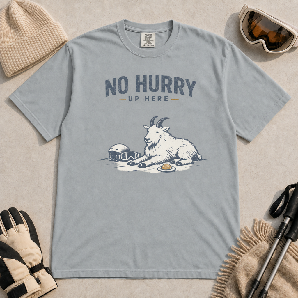 Granite gray t-shirt featuring a mountain goat relaxing in the snow with a helmet, goggles and snack beneath the words “No Hurry Up Here.”