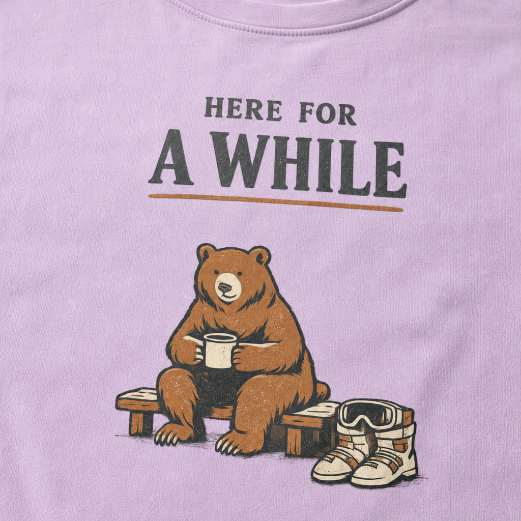 Close-up of an orchid purple t-shirt featuring a bear holding a mug beside ski boots and goggles with the phrase “Here for a While.”