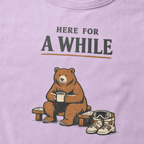 Close-up of an orchid purple t-shirt featuring a bear holding a mug beside ski boots and goggles with the phrase “Here for a While.”