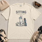 Ivory white t-shirt featuring a goggle-wearing rabbit resting on a bench beside a mug and snowboard with “Sitting This One Out” text.