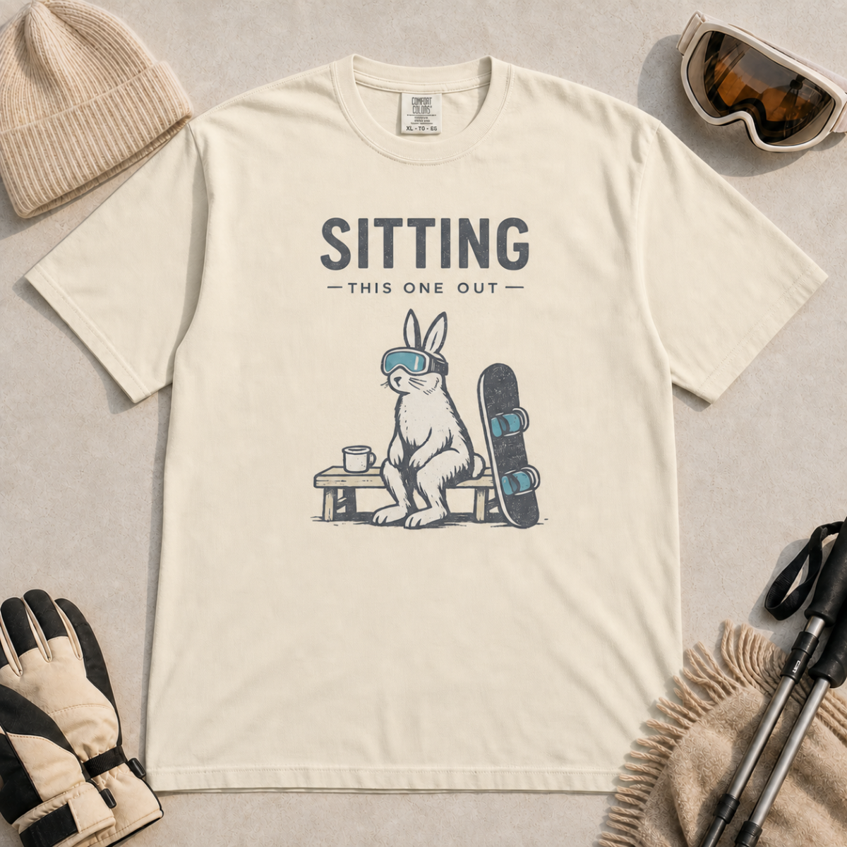 Ivory white t-shirt featuring a goggle-wearing rabbit resting on a bench beside a mug and snowboard with “Sitting This One Out” text.
