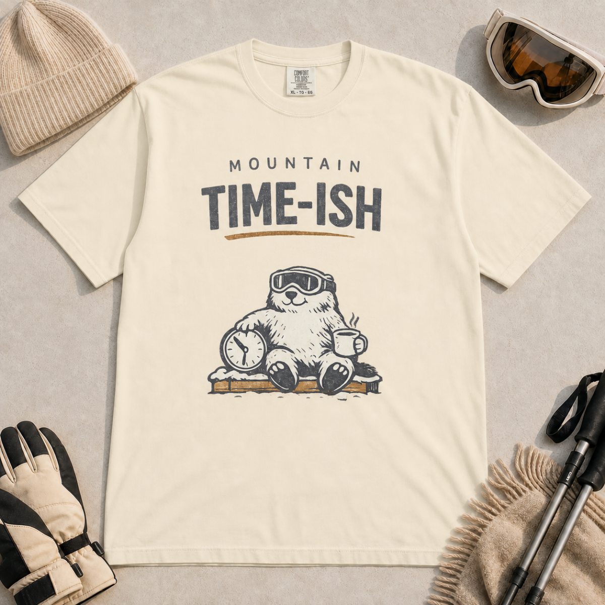 Ivory white t-shirt featuring a relaxed marmot in ski goggles holding a clock and steaming mug beneath the words “Mountain Time-ish.”
