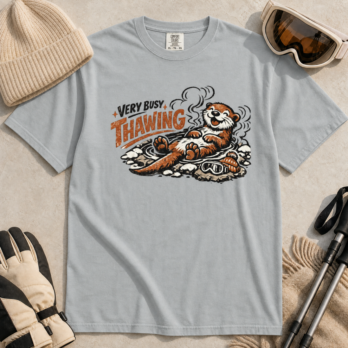 Granite gray t-shirt featuring a smiling otter thawing in a rocky hot spring beside ski goggles and a beanie.
