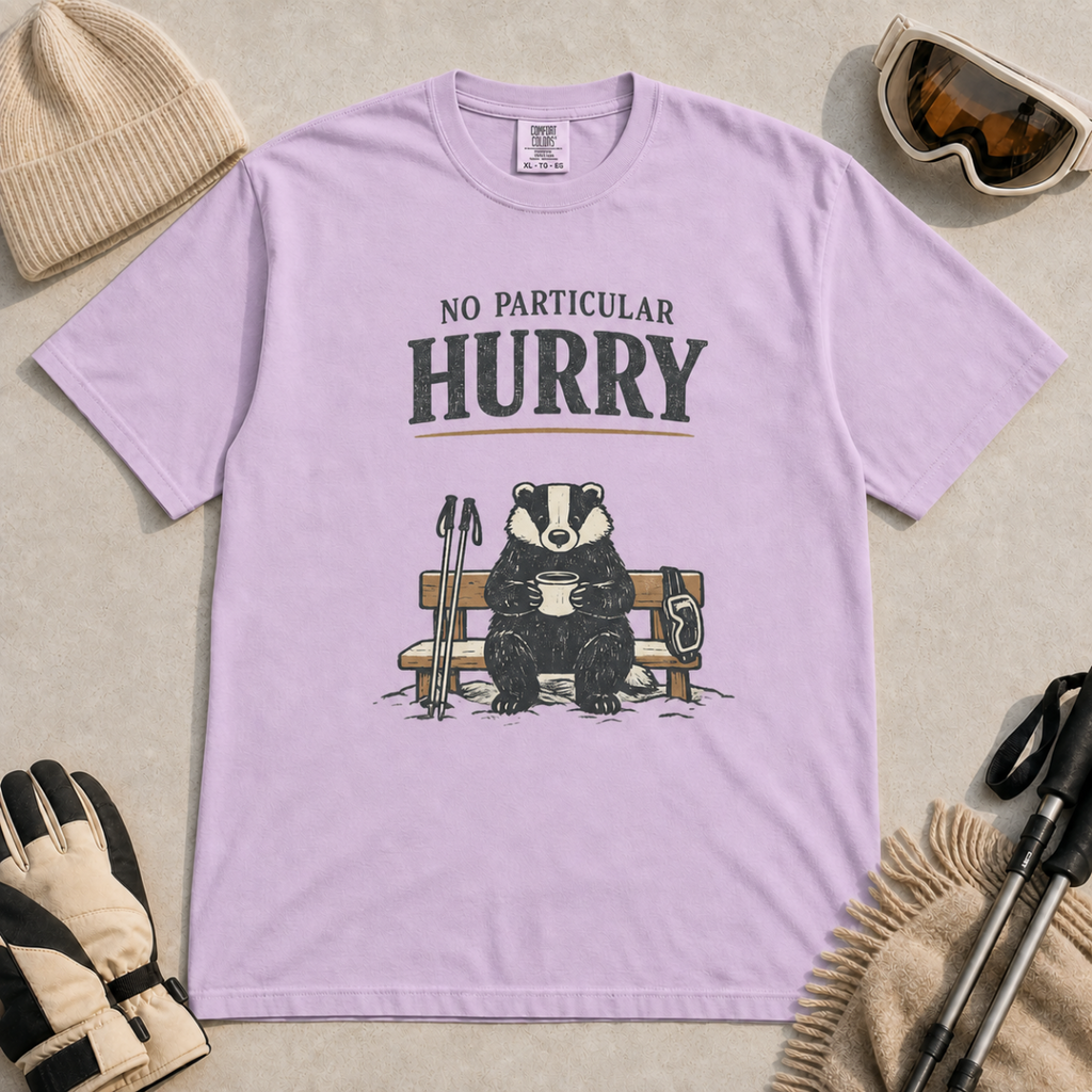 Orchid purple t-shirt featuring a badger relaxing on a snowy bench with a mug, ski poles and goggles beneath the words “No Particular Hurry.”