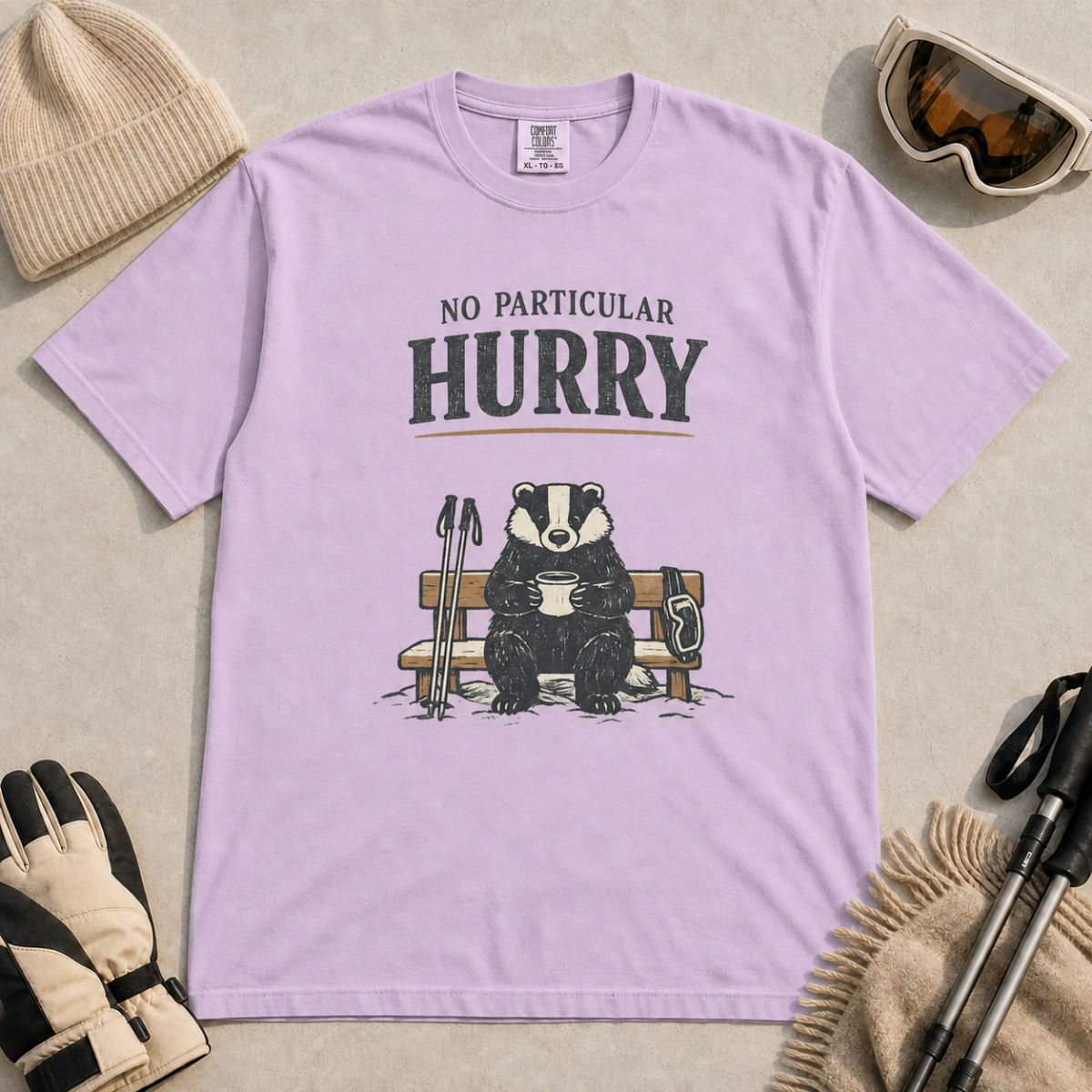 Orchid purple t-shirt featuring a badger relaxing on a snowy bench with a mug, ski poles and goggles beneath the words “No Particular Hurry.”