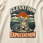 Close-up of a t-shirt design featuring a small mountain rodent holding a mug beside ski goggles, with sunset peaks and the phrase “Elevation Without Expectation."