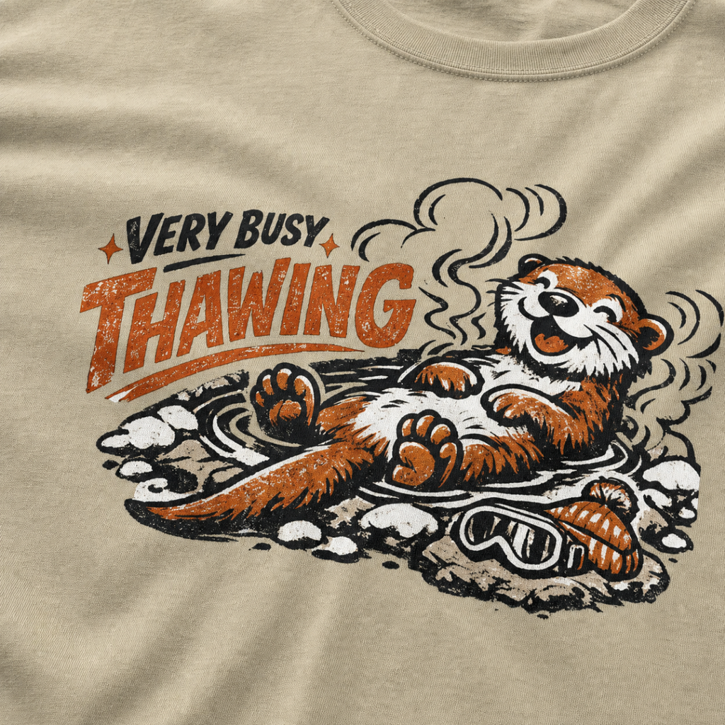 Close-up of a khaki t-shirt featuring a relaxed otter soaking in a steaming hot spring beneath “Very Busy Thawing” text.