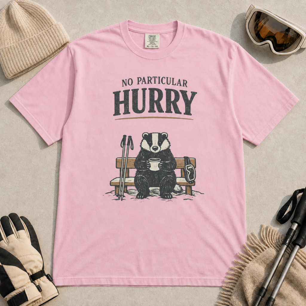Blossom pink t-shirt featuring a badger relaxing on a snowy bench with a mug, ski poles and goggles beneath the words “No Particular Hurry.”