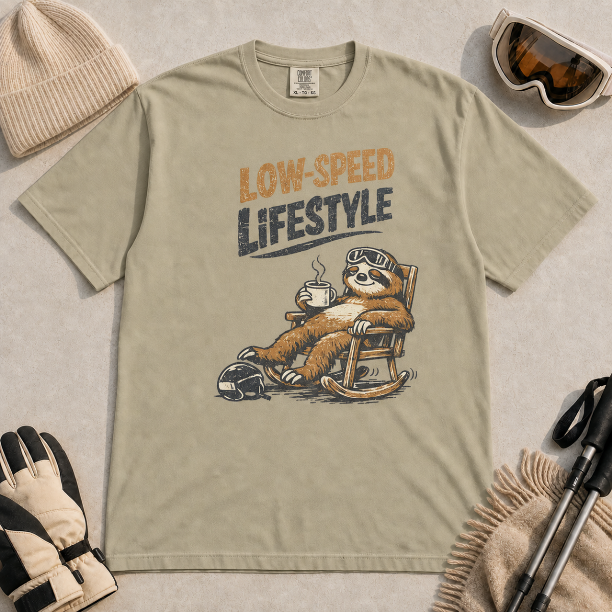 Khaki t-shirt featuring a relaxed sloth in ski goggles rocking in a chair with a hot drink beneath the words “Low-Speed Lifestyle.”