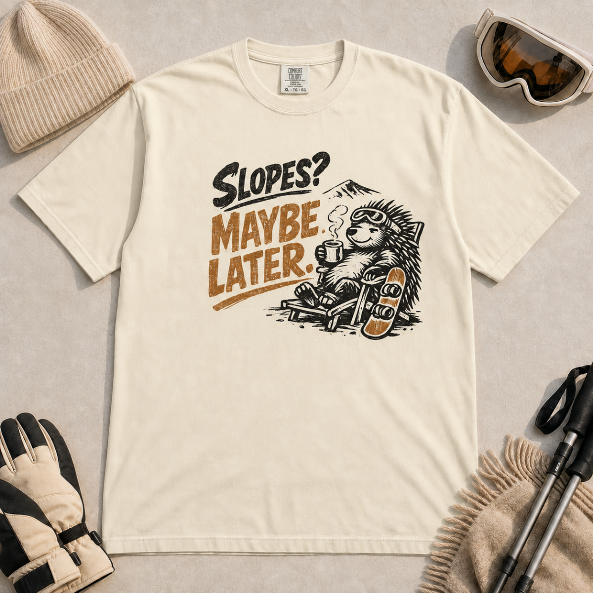 Ivory white t-shirt featuring a relaxed hedgehog in a chair with a hot drink and snowboard beneath “Slopes? Maybe Later.” text.