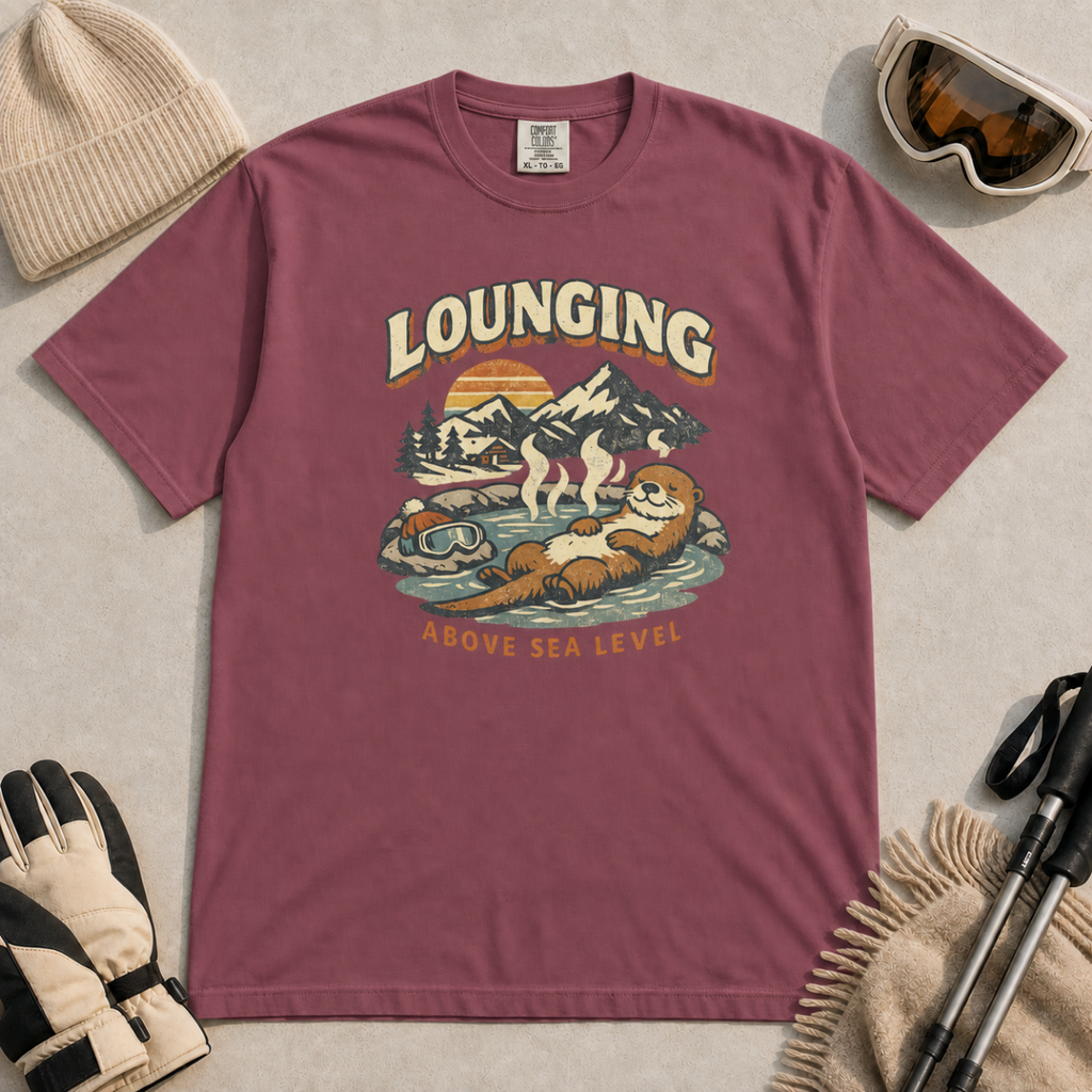 Brick red t-shirt featuring an otter relaxing in a steaming mountain hot spring beneath the words “Lounging Above Sea Level.”