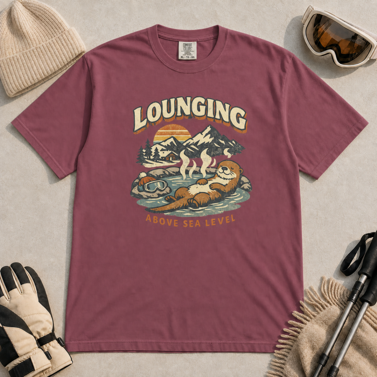 Brick red t-shirt featuring an otter relaxing in a steaming mountain hot spring beneath the words “Lounging Above Sea Level.”