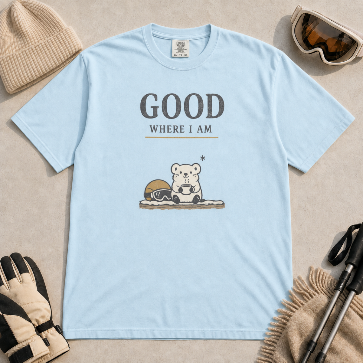 Chambray blue t-shirt featuring a small white bear holding a mug beside ski goggles with the phrase “Good Where I Am.”